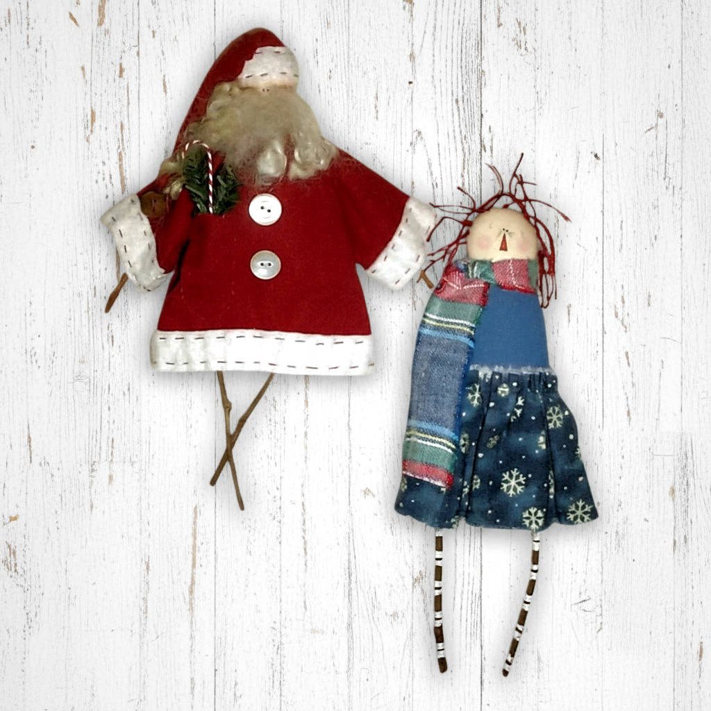 Two handmade dolls, one dressed as Santa Claus and the other in a blue dress with floral patterns, on a white wooden background.