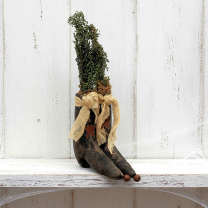Decorative pair of rustic witch boots with dried plants and a bow on a white wooden surface.