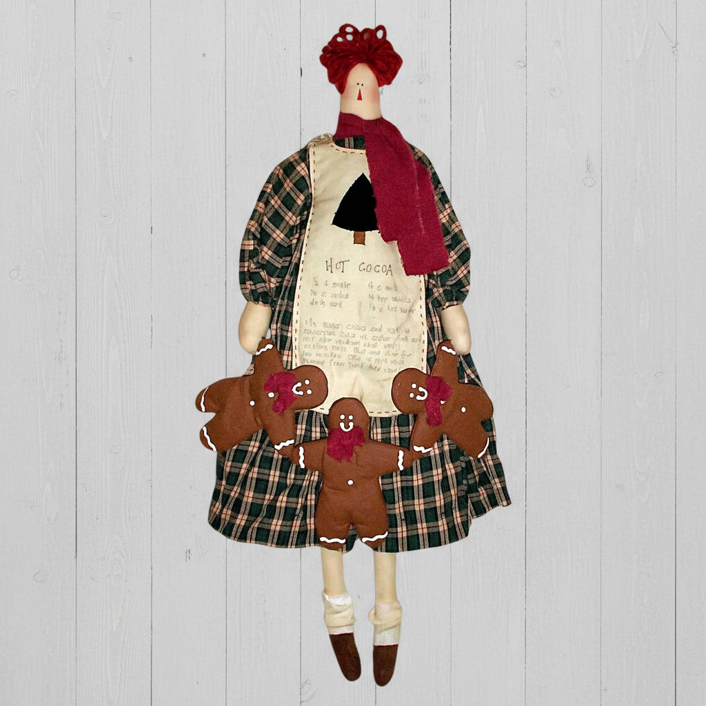 Decorative rag doll with plaid skirt and red hair, surrounded by gingerbread cookies on a wooden background.