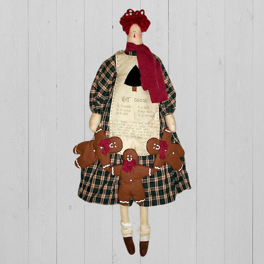 Decorative rag doll with plaid skirt and red hair, surrounded by gingerbread cookies on a wooden background.