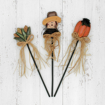Three decorative sticks with a scarecrow, corn, and pumpkin on a wooden background
