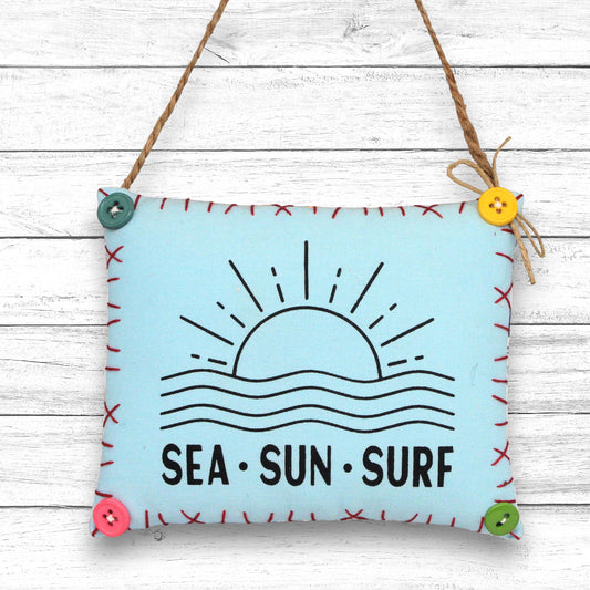 Decorative sign ornament with 'Sea Sun Surf' text and sun and wave design on a light blue background, hanging on a wooden surface.
