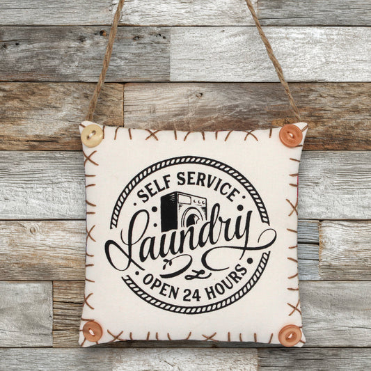Fabric sign ornament with 'Self Service Laundry Open 24 Hours' text on a wooden background