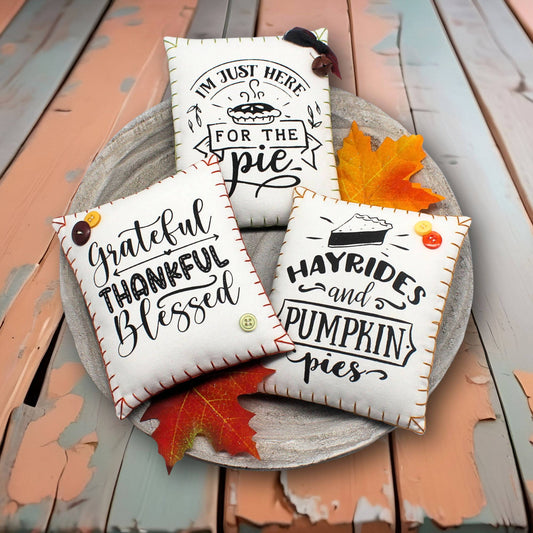 Three decorative pillows with autumn-themed sayings on a wooden surface with fall leaves.