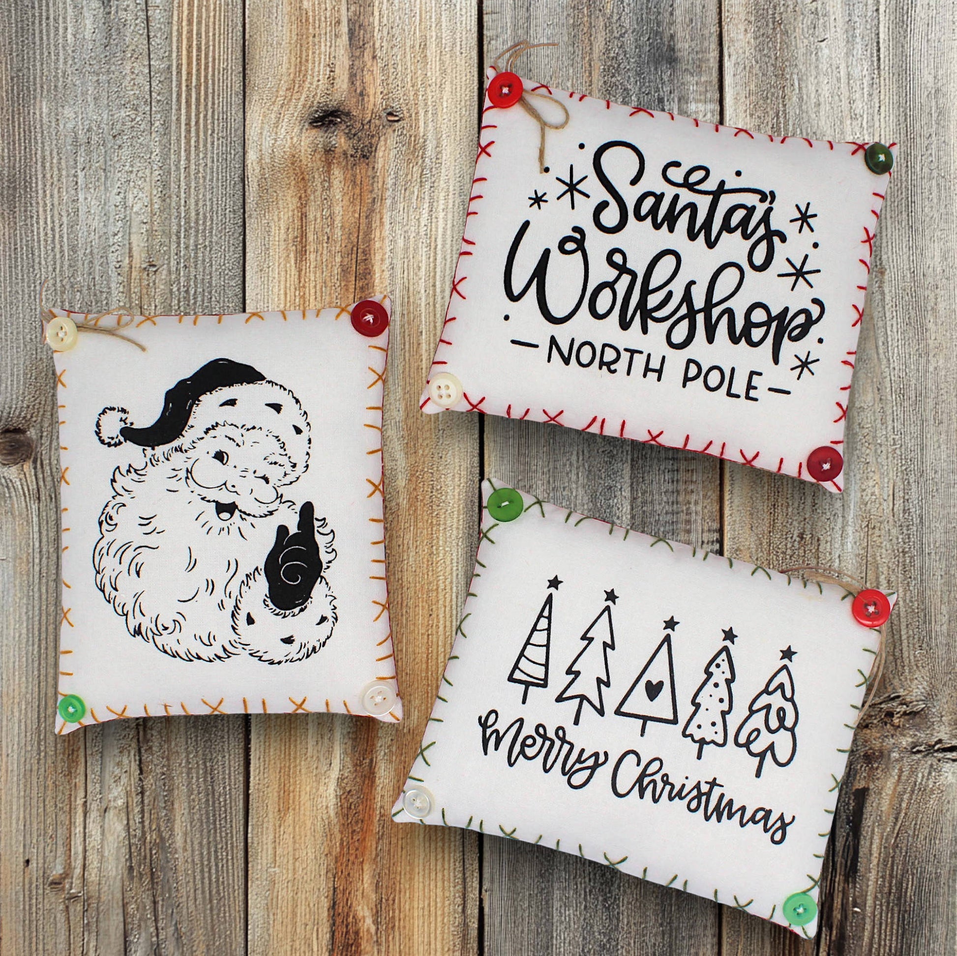 Three Christmas-themed mini pillows on a wooden surface with 'Santa's Workshop North Pole' and 'Merry Christmas' text.
