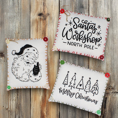 Three Christmas-themed mini pillows on a wooden surface with 'Santa's Workshop North Pole' and 'Merry Christmas' text.