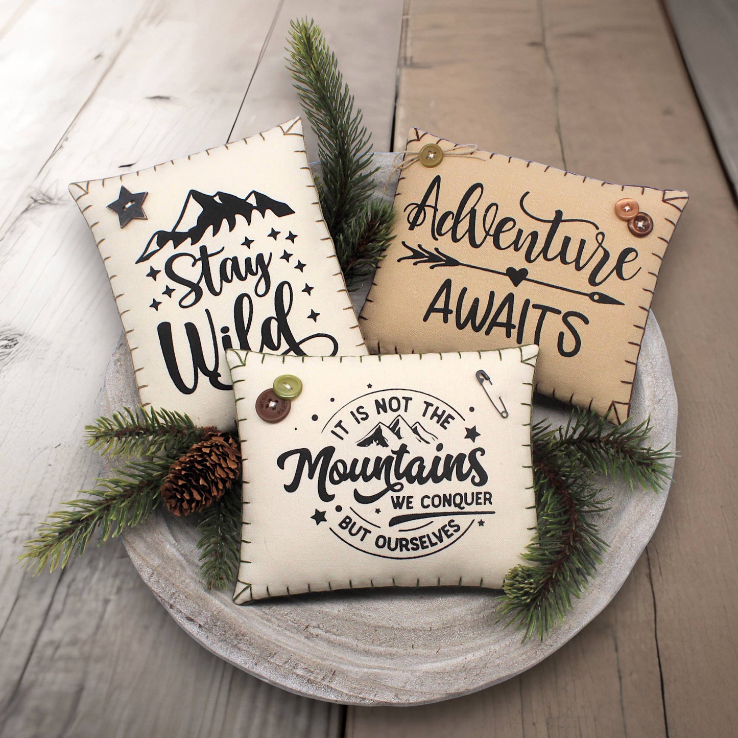 Three decorative pillows with motivational phrases on a wooden surface with pine branches.