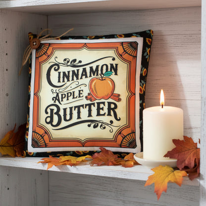 Decorative pillow with 'Cinnamon Apple Butter' text on a shelf with leaves and a candle.