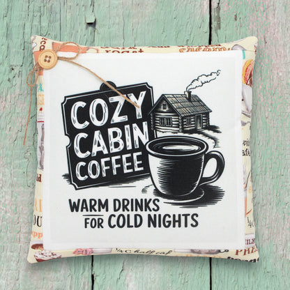 Decorative pillow with 'Cozy Cabin Coffee' text and a cup of coffee graphic on a wooden surface.