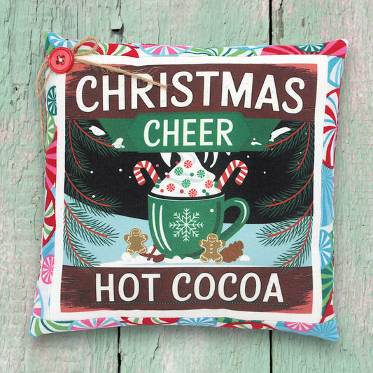 Decorative pillow with 'Christmas Cheer Hot Cocoa' text and festive design on a wooden surface.