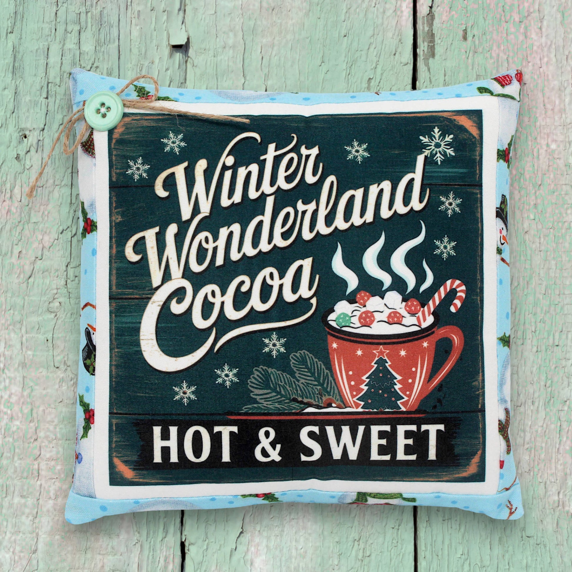Decorative pillow with 'Winter Wonderland Cocoa Hot & Sweet' text and hot chocolate illustration on a wooden background.
