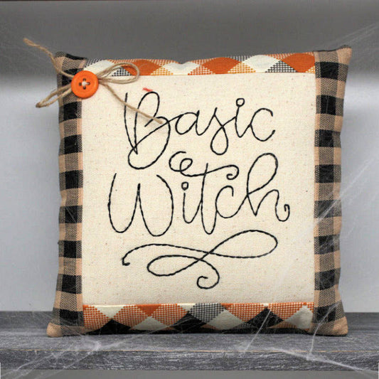 Decorative pillow with 'Basic Witch' text and plaid pattern on a neutral background